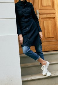 Vestito camicia blu navy con due tasche sul petto, abbinato a jeans in denim chiaro arrotolati sui polsini e sneakers bianche con lacci.