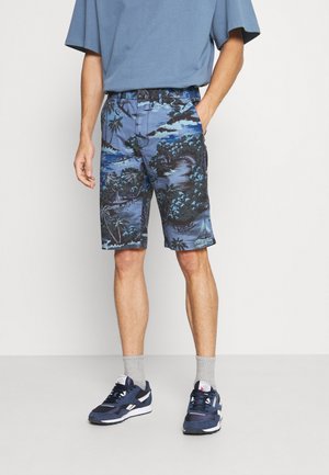 Mann trägt blaue Shorts mit tropischem Muster, blaues T-Shirt, graue Socken und marineblaue Turnschuhe und steht vor einem schlichten hellen Hintergrund.