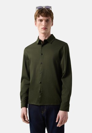 Ung mand iført mørkegrøn langærmet skjorte med knapper og mørkeblå jeans med solbriller hvilende på hovedet, neutral baggrund.