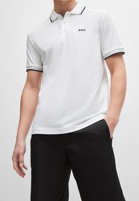 Polo shirt blanc à manches courtes, avec une accent noir au col et aux bords des manches, présentant un petit logo noir "BOSS" sur la poche gauche de la poitrine.