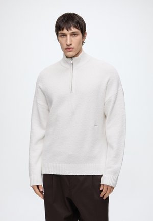 Young man wearing a white textured half-zip sweater and dark pants against a plain light background.