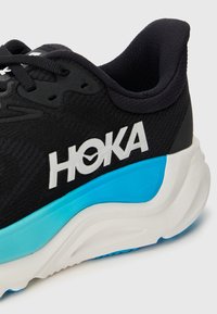 Schwarzer Laufschuh mit Netzobermaterial, ausgeprägtem weißen Logo und auffälligem türkisfarbenen Akzent auf der Zwischensohle. Verfügt über eine strukturierte Sohle für besseren Grip.