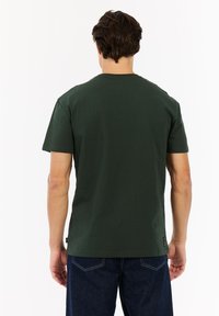 Man met kort bruin haar, gekleed in een donkergroene T-shirt met korte mouwen en donkerblauwe spijkerbroeken, met zijn rug naar de camera gericht tegen een witte achtergrond.