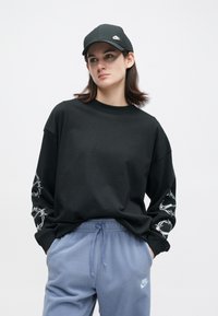 Mladý človek s čiernou čiapkou Nike, čiernym svetrom so vzorom ostnatého drôtu na rukávoch a svetlomodrými teplákmi Nike, so stoletými rukami v vreckách.