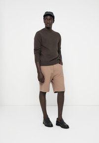 Brun stickad tröja, beige randiga shorts och svarta slip-on skor. Modell bär en svart keps och står framför en helt vit bakgrund.