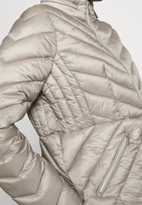 Jasnobeżowa pikowana kurtka z wysokim kołnierzem. Posiada wzór w romby, diagonalne szwy oraz zapinaną na zamek kieszeń boczną. Gładka i błyszcząca tekstura.