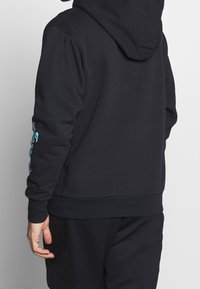 Sudadera con capucha negra y mangas largas. Presenta un tejido texturizado y un diseño gráfico azul a lo largo del brazo izquierdo. Corte simple y relajado.