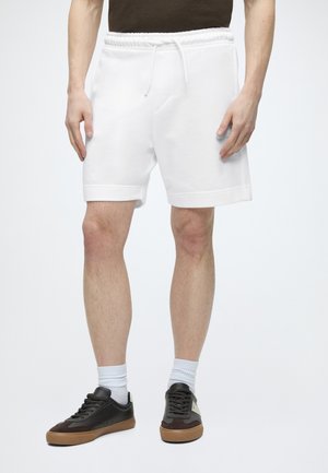 Muž, ktorý má na sebe biele šortky so šnúrkou, biele ponožky a čierne nízke tenisky s hnedými podrážkami, stojí na jednoduchej bielej pozadí.