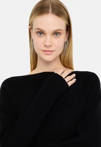 Maglione nero a coste con una vestibilità comoda, caratterizzato da maniche lunghe. Il modello indossa orecchini lunghi in argento e ha i capelli biondi lisci.