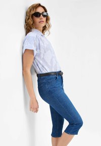 Camicetta a maniche corte azzurro chiaro con motivo floreale, abbinata a jeans in denim blu scuro a lunghezza ginocchio e una cintura nera. Il modello indossa occhiali da sole scuri.
