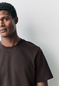 T-shirt en coton marron avec un col rond et des manches courtes. Texture lisse, détails de coutures minimaux et coupe ajustée.