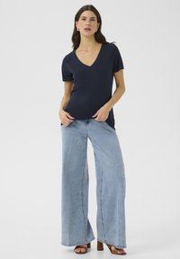 Camiseta de cuello en V de color marino, confeccionada en una tela suave, combinada con jeans de pierna ancha de color azul claro. El modelo lleva sandalias de tacón marrón. Un look simple y casual.