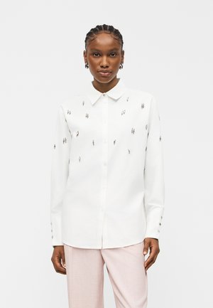 Žena má na sebe bielu košeľu so zapínaním na gombíky a malými striebornými ozdobami a svetloružové nohavice, stojí pred jednoduchým svetlým pozadím.