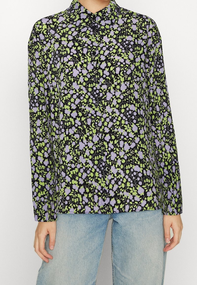 Chemise à manches longues boutonnée avec fond noir et motif floral vert et violet, portée avec un jean bleu clair.