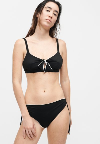 Γυναίκα με βρεγμένα μαλλιά, που φοράει μαύρο μπικίνι με δέσιμο μπροστά στο επάνω μέρος και δέσιμο στα πλάγια στο κάτω μέρος, σε απλό λευκό φόντο.