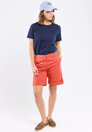 Femme portant un t-shirt bleu marine, un short corail retroussé, des chaussures bateau marron et une casquette de baseball à rayures bleu clair, debout avec une main dans la poche.