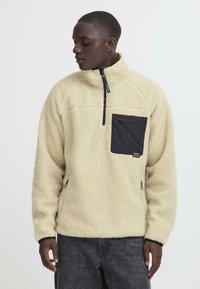 Fleece pullover in cream with a quarter zipper, textured fabric, and a black chest pocket featuring a zip closure.