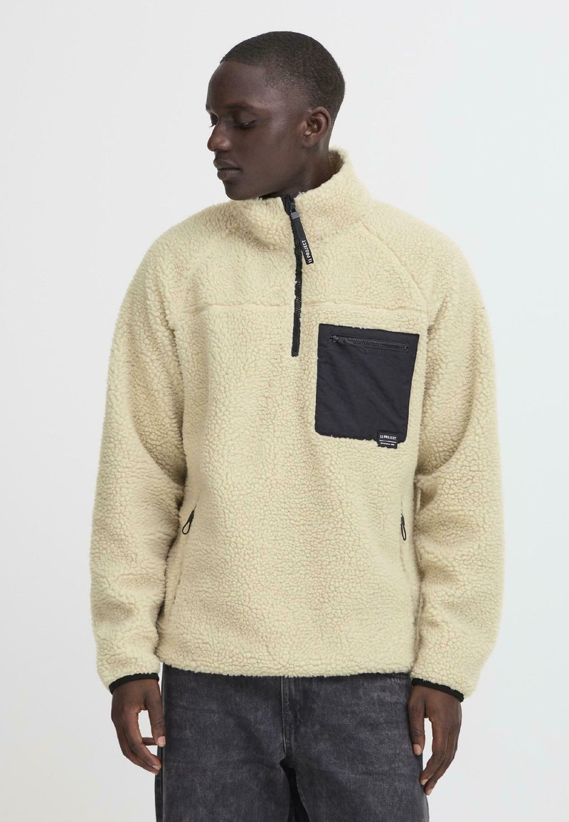 Fleece pullover in cream with a quarter zipper, textured fabric, and a black chest pocket featuring a zip closure.