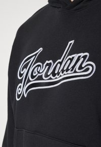 Gros plan sur un sweat à capuche noir avec le logo "Jordan" en cursive blanche brodé sur la poitrine.