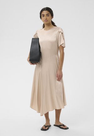 Mujer con vestido midi de satén beige sosteniendo un bolso negro, calzando sandalias negras, de pie frente a un fondo blanco liso.