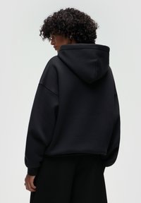 Felpa nera con vestibilità rilassata, cappuccio oversize e cuciture delle spalle abbassate. Texture morbida e polsini a coste. Indossata con pantaloni neri a vita alta.