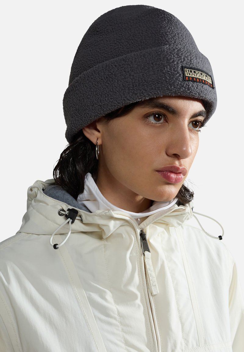 Gorro de forro polar gris con un diseño acanalado y parche de logo. Presenta un ajuste ceñido, acompañado de una chaqueta blanca con capucha y cierre de cremallera.