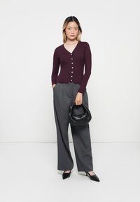 Gilet long bordeaux à manches longues avec bordure en dentelle et fermeture par boutons associé à un pantalon gris taille haute et ample, accompagné d'un sac à main noir.