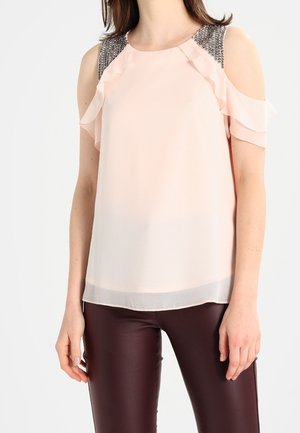 Kvinde iført en lyserød cold-shoulder bluse med flæseærmer og dekorerede skuldre, kombineret med mørkebrune bukser.