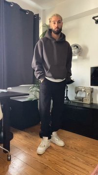 Black cotton pants, gray hoodie with a front pocket, white t-shirt underneath, and white sneakers, standing in a room with wooden flooring.