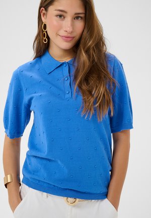Femme aux longs cheveux bruns portant un polo bleu texturé à manches courtes, un pantalon blanc avec une ceinture tressée, des boucles d'oreilles en or et un bracelet en or.