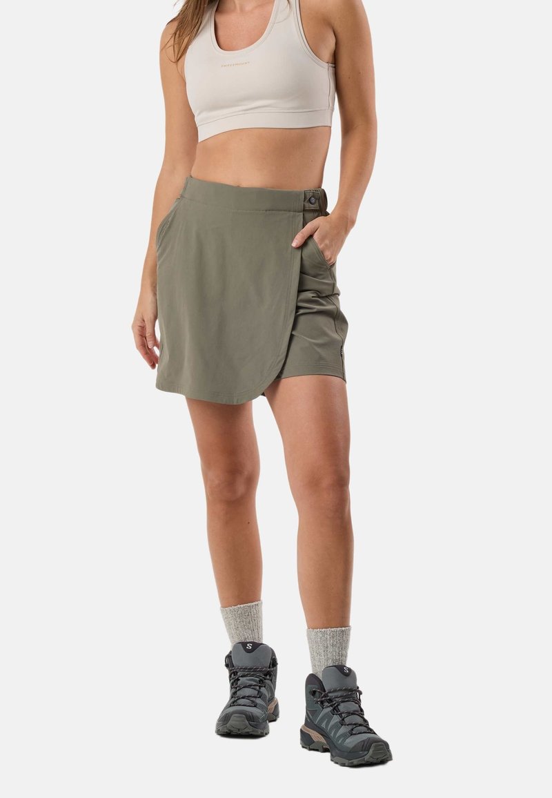Femme portant une jupe portefeuille kaki avec un short en dessous, un soutien-gorge de sport beige, des chaussettes grises et des chaussures de randonnée noires, debout avec une main dans la poche.