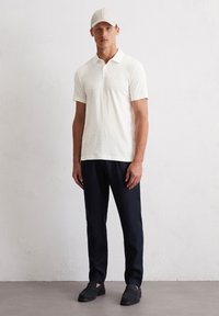 Lichtbeige poloshirt met een klassieke kraag, gecombineerd met donkerblauwe broek. De outfit wordt aangevuld met een bijpassende beige pet en donkere instappers.