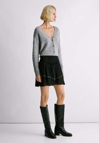 Cardigan grigio corto con bottoni, gonna nera svasata con strati a balze e stivali neri in pelle alti fino al ginocchio con tacco a blocco.