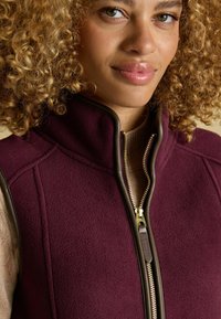 Weinroter Fleece-Reißverschlussweste mit braunen Akzenten, ausgestattet mit einem hohen Kragen und goldenen Reißverschlussdetails. Glatte Textur mit Seitenstichen.