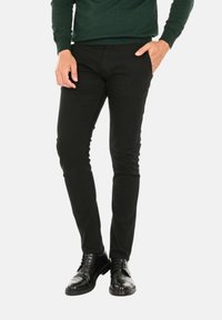 Pantaloni neri slim-fit in denim, caratterizzati da una texture liscia. Abbinati a scarpe eleganti nere lucide e a un maglione verde scuro.