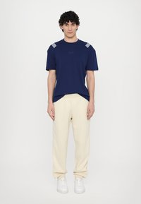T-shirt bleu marine à manches courtes avec des accents rayés sur les épaules, associé à un pantalon de jogging crème et des baskets blanches. Style simple et décontracté.