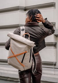 Beige rolltop rugzak gemaakt van duurzaam materiaal, met een oranje riem, zwarte ritsen en een minimalistisch ontwerp met een gestructureerde afwerking.