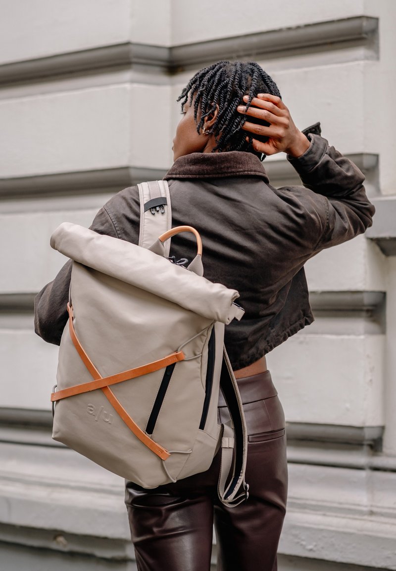 Beige rolltop rugzak gemaakt van duurzaam materiaal, met een oranje riem, zwarte ritsen en een minimalistisch ontwerp met een gestructureerde afwerking.