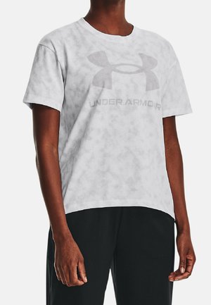 Osoba ubrana w jasnoszary t-shirt Under Armour z dużym logo i napisem, w zestawieniu z czarnymi spodniami, stojąca na jednolitym tle.