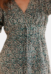 Kleid mit tiefem V-Ausschnitt, mit einem floralem Muster in Teal, Creme und Schwarz. Weicher Stoff mit gerafften Nähten und lockeren Ärmeln.