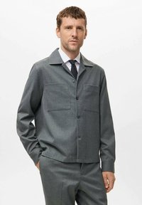 Chaqueta gris texturizada con collar, dos bolsillos en el pecho y corte recto. Llevada sobre una camisa blanca y una corbata negra. Botones en la parte frontal.