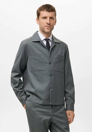 Chaqueta gris texturizada con collar, dos bolsillos en el pecho y corte recto. Llevada sobre una camisa blanca y una corbata negra. Botones en la parte frontal.