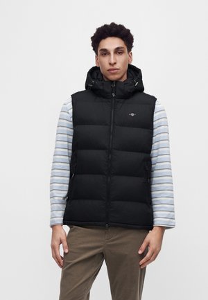 Jeune homme aux cheveux bouclés porte une veste matelassée noire avec capuche sur une chemise légère à rayures manches longues et un pantalon marron, debout devant un fond blanc.