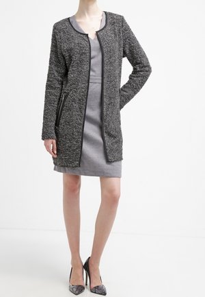Femme portant un manteau gris texturé mi-long par-dessus une robe grise cintrée et des chaussures à talons hauts à motifs, debout, une main sur la hanche.