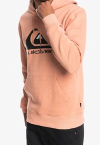 Vaaleanpunainen puuvillapaitapusero, jossa on etutasku, nyörillinen huppu ja musta logografiikka rinnassa. Pehmeä rakenne, ribbikuvioidut hihansuut ja helma.