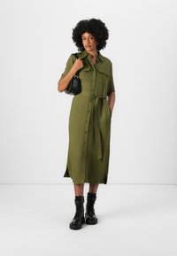 Vestito verde oliva con bottoni, maniche corte, due tasche sul petto, una cintura legata in vita e spacchi laterali. Indossato con stivaletti neri e borsa.