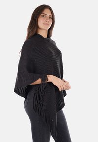 Poncho nero lavorato a maglia con texture a coste, caratterizzato da frange lungo l'orlo e maniche aperte, drappeggiato sopra le spalle.