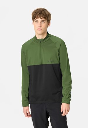 Giovane uomo che indossa una giacca outdoor verde e nera con mezza cerniera, in piedi davanti a uno sfondo chiaro e uniforme.