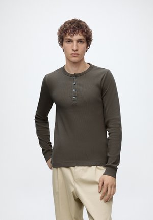 Jeune homme aux cheveux bouclés portant une chemise Henley à manches longues vert foncé et un pantalon beige, debout devant un fond clair uni.