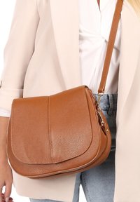 Sac en cuir marron à bandoulière avec un design à rabat, surface texturée, garnitures dorées et une bandoulière ajustable. Style compact et pratique.